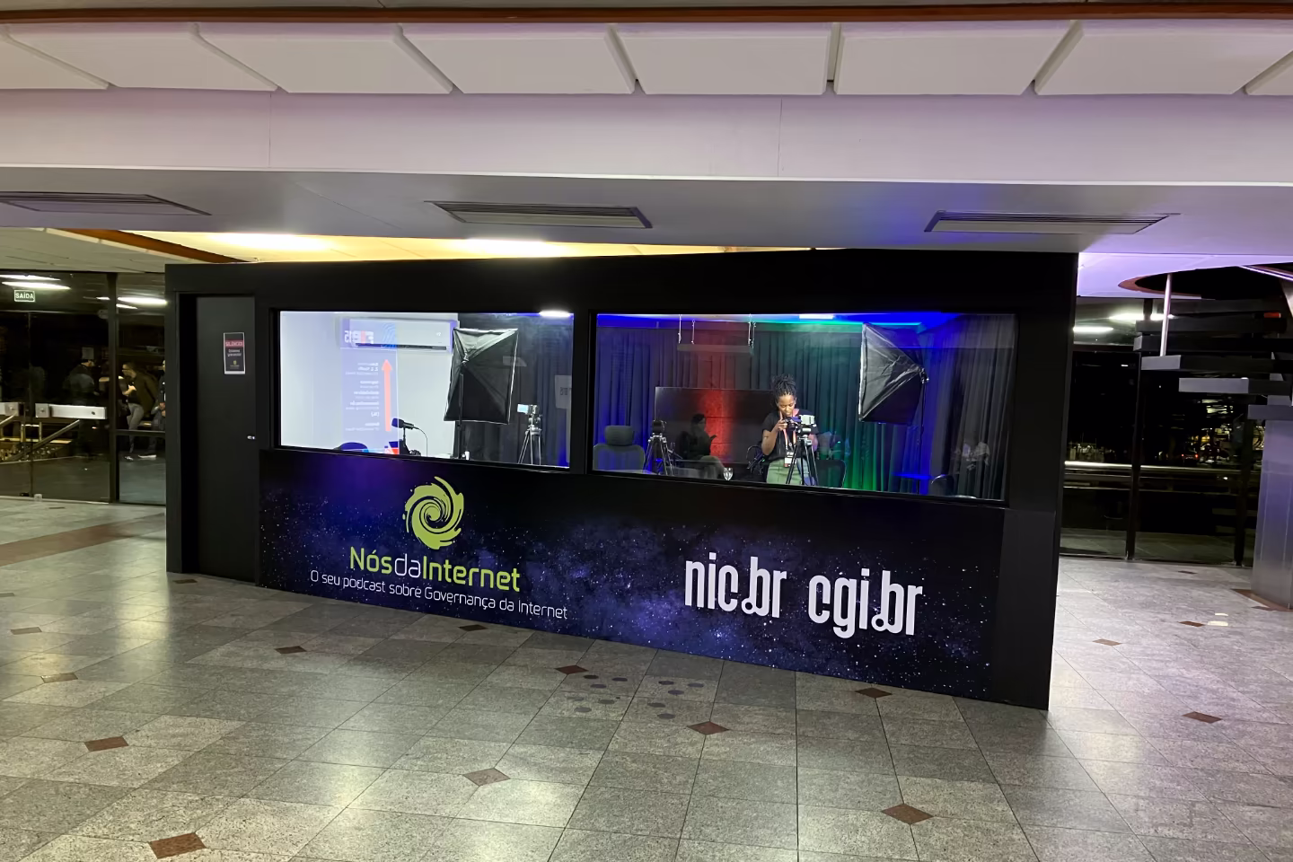 Estúdio com janelas amplas, envelopado com os logos do podcast Nós da Internet, CGI.br e NIC.br.
