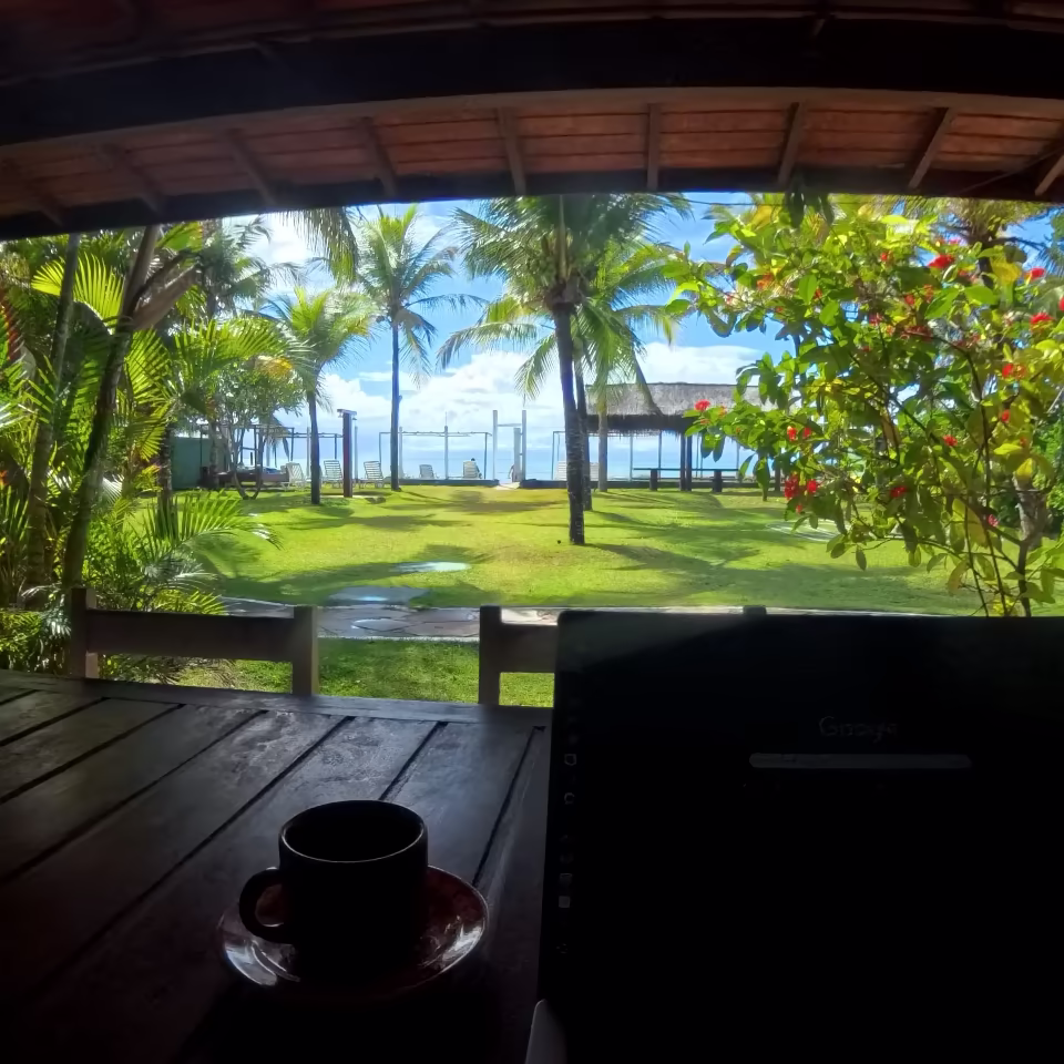 Paisagem com gramado e árvores, em frente ao mar, com uma xícara em cima da mesa em primeiro plano.