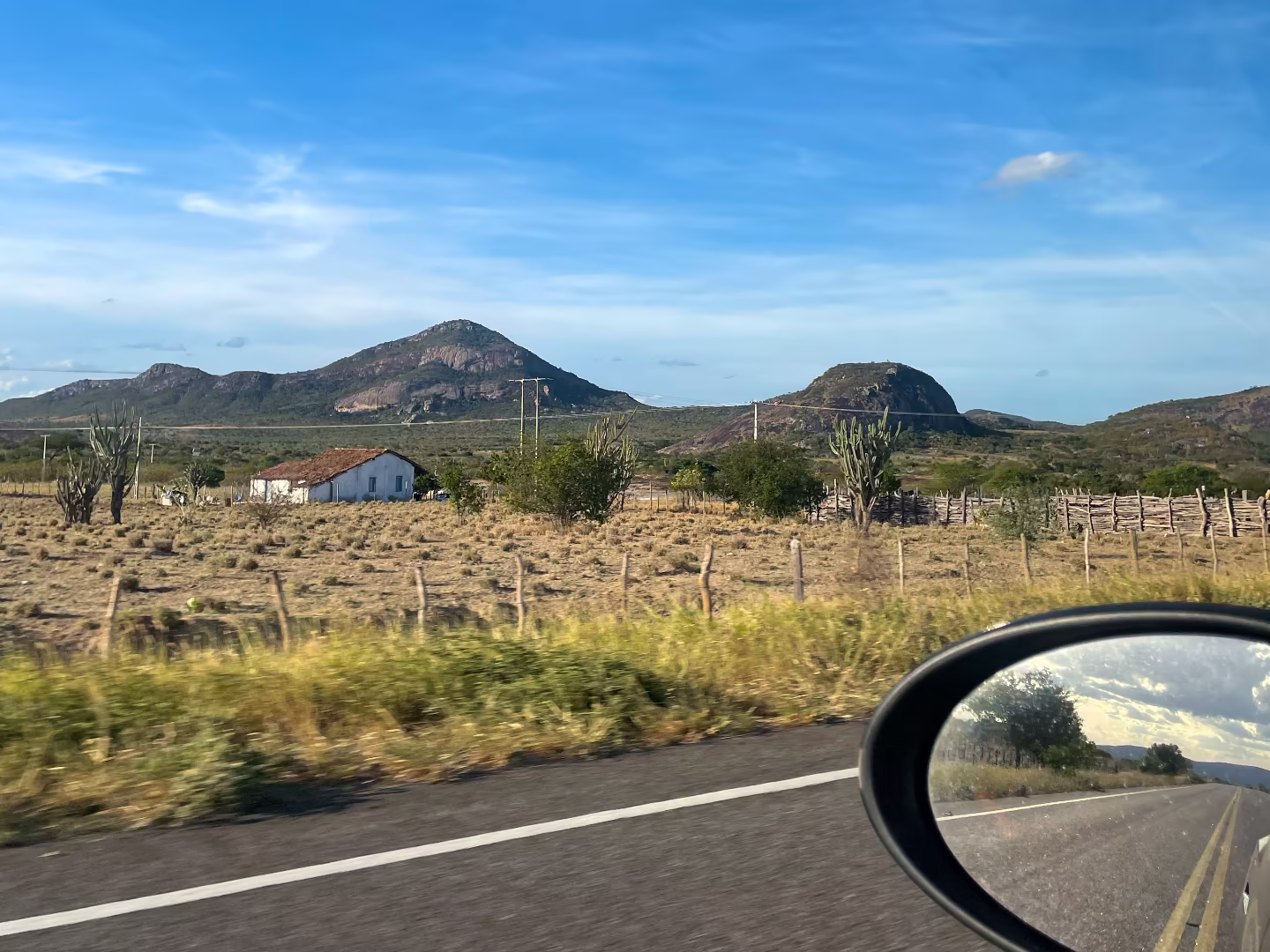 Paisagem árida, com céu azul, a bordo de um carro; retrovisor parcialmente visível no canto inferior direito.