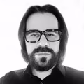 Homem branco, de óculos de grau, barba preta e cabelo comprido, preto e liso. Foto em preto e branco.