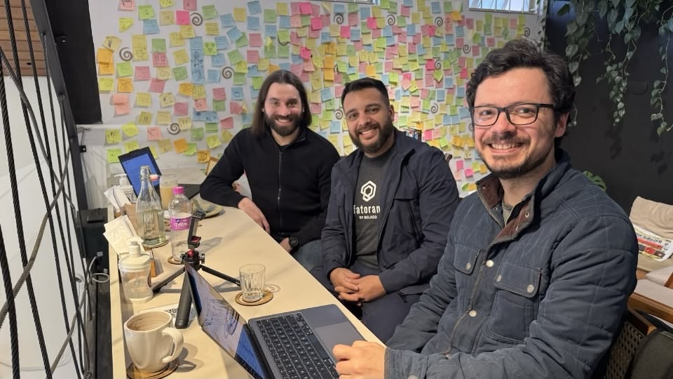 Três homens brancos sentados em frente a uma bancada, sorrindo, com uma parede cheia de post-its atrás.