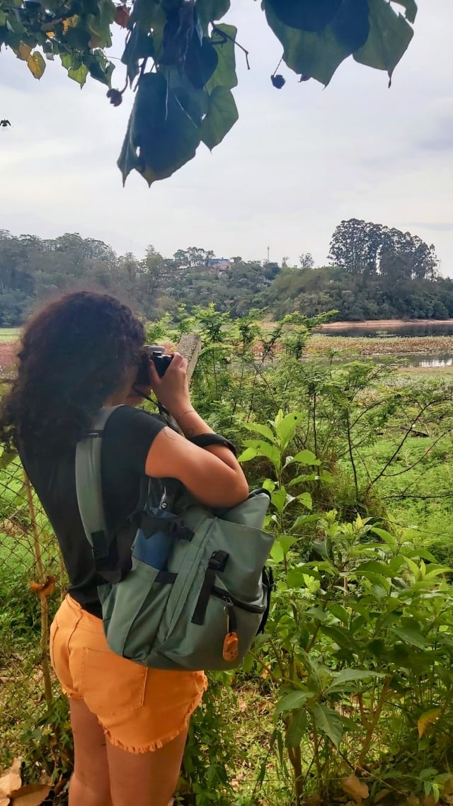 Mulher de costas, fotografando numa área verde, com uma mochila em destaque, pendurada em um dos ombros.