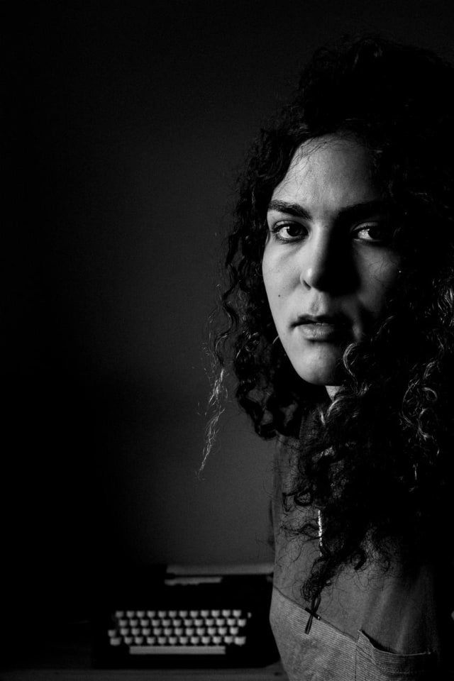 Foto em preto e branco de uma mulher, olhando de lado, com uma máquina de escrever ao fundo.