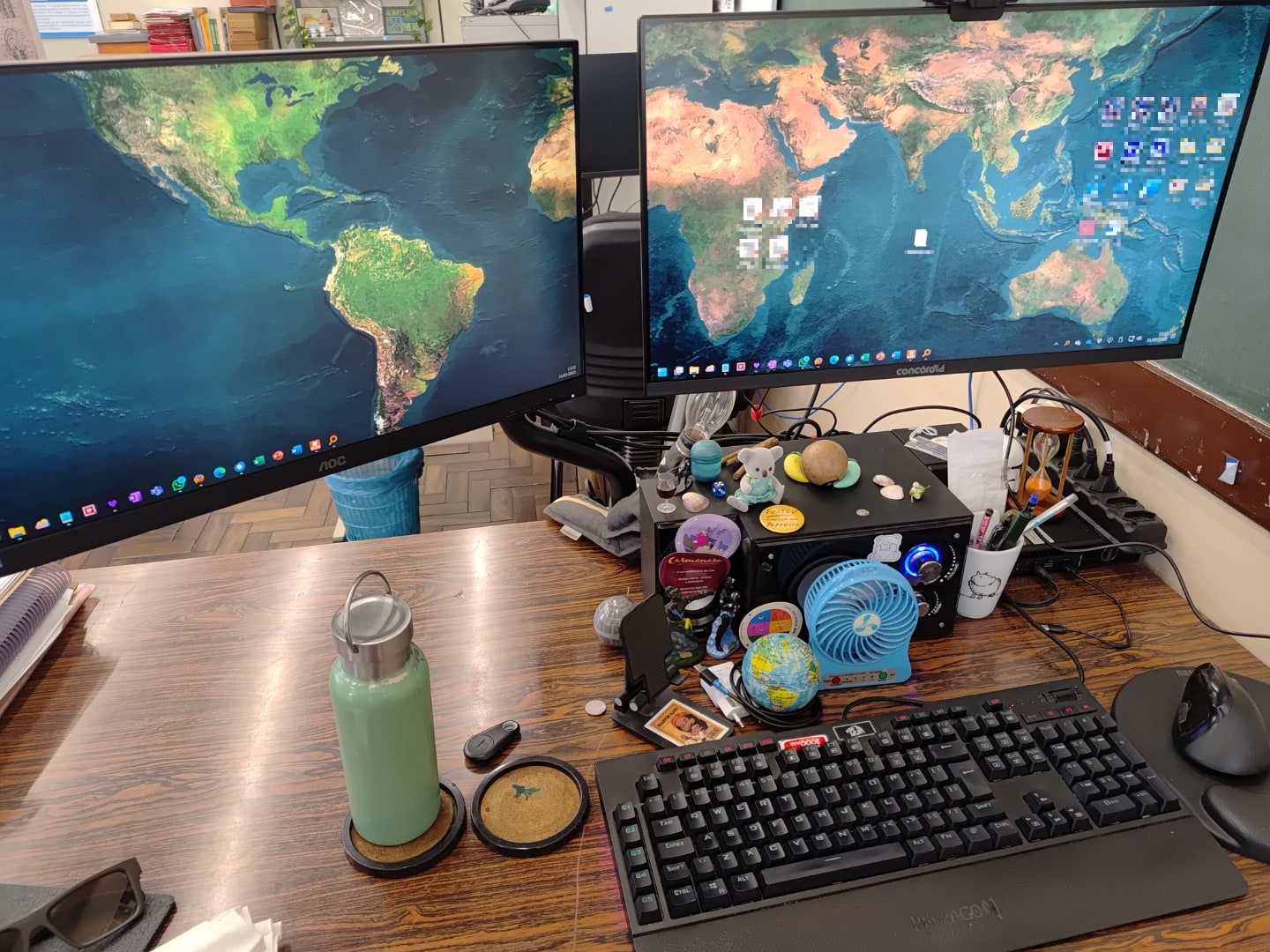 Mesa com teclado, mouse, vários objetos e dois monitores lado a lado, com papel de parede do globo terrestre.