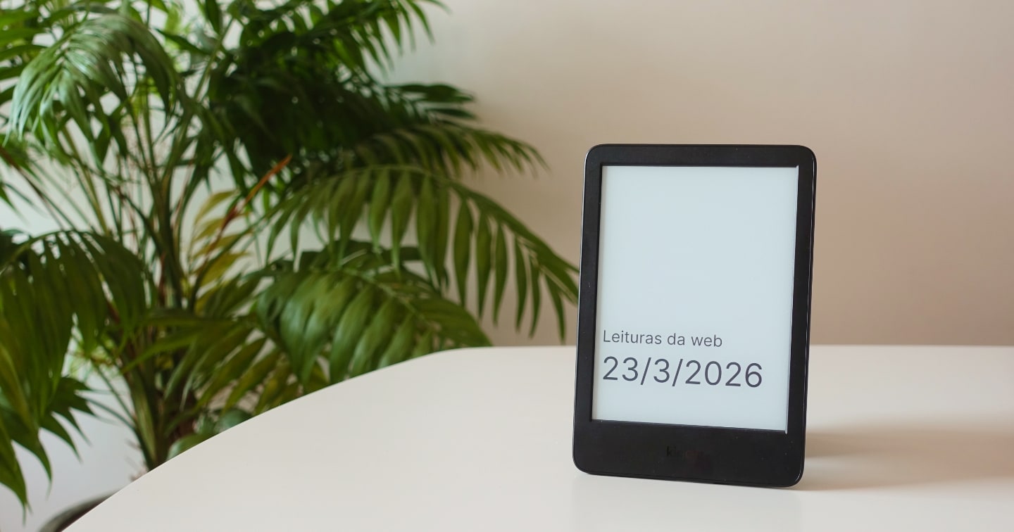 Kindle sobre uma mesa branca, exibindo a capa de um livro onde se lê “Leituras da web 23/3/2026”. Ao fundo, uma palmeira pequena.
