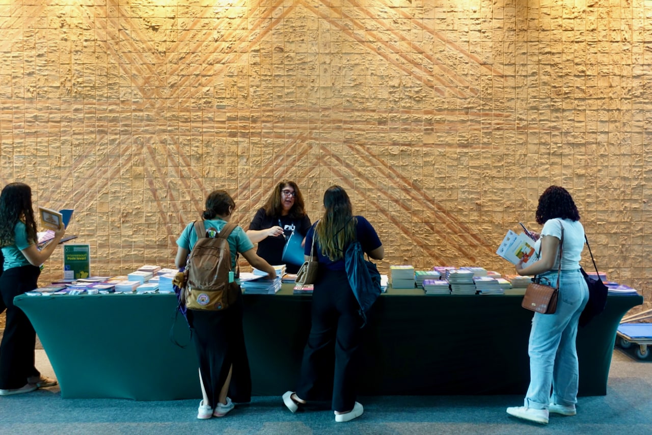 A long table full of books on top, in front of a textured beige wall, with people around it.