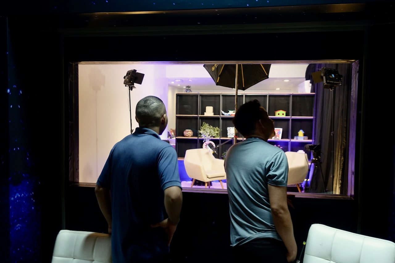 Two men observing a studio that looks like a living room through a glass window.
