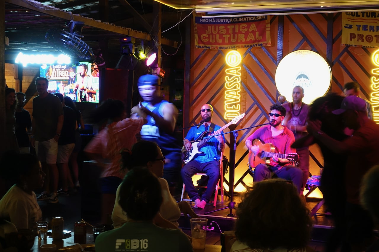 Stage with musicians playing and, in front, silhouettes of some people in motion, dancing.