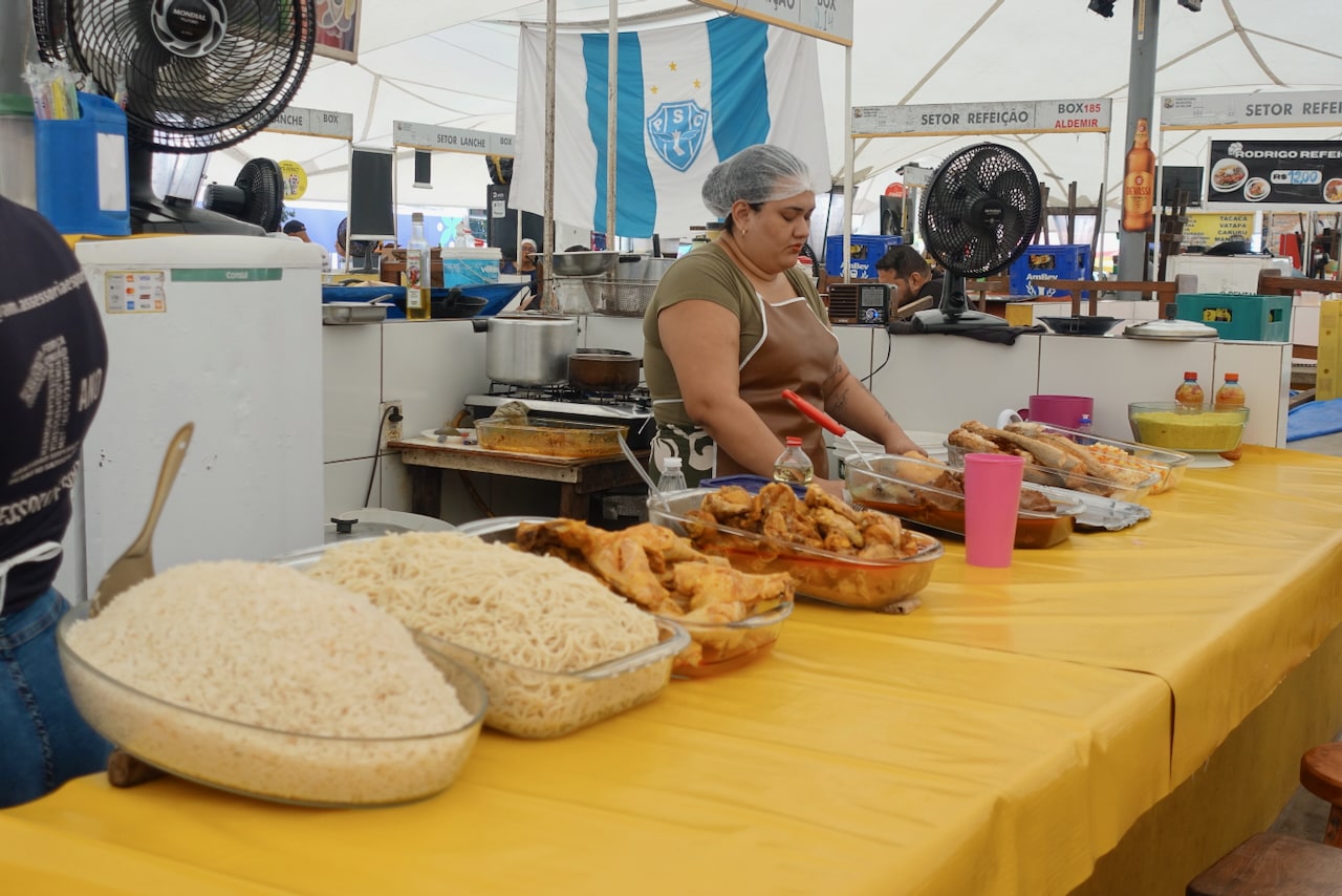 Market stall with large bowls of cooked food, like rice and meats, and a woman behind the counter wearing an apron.