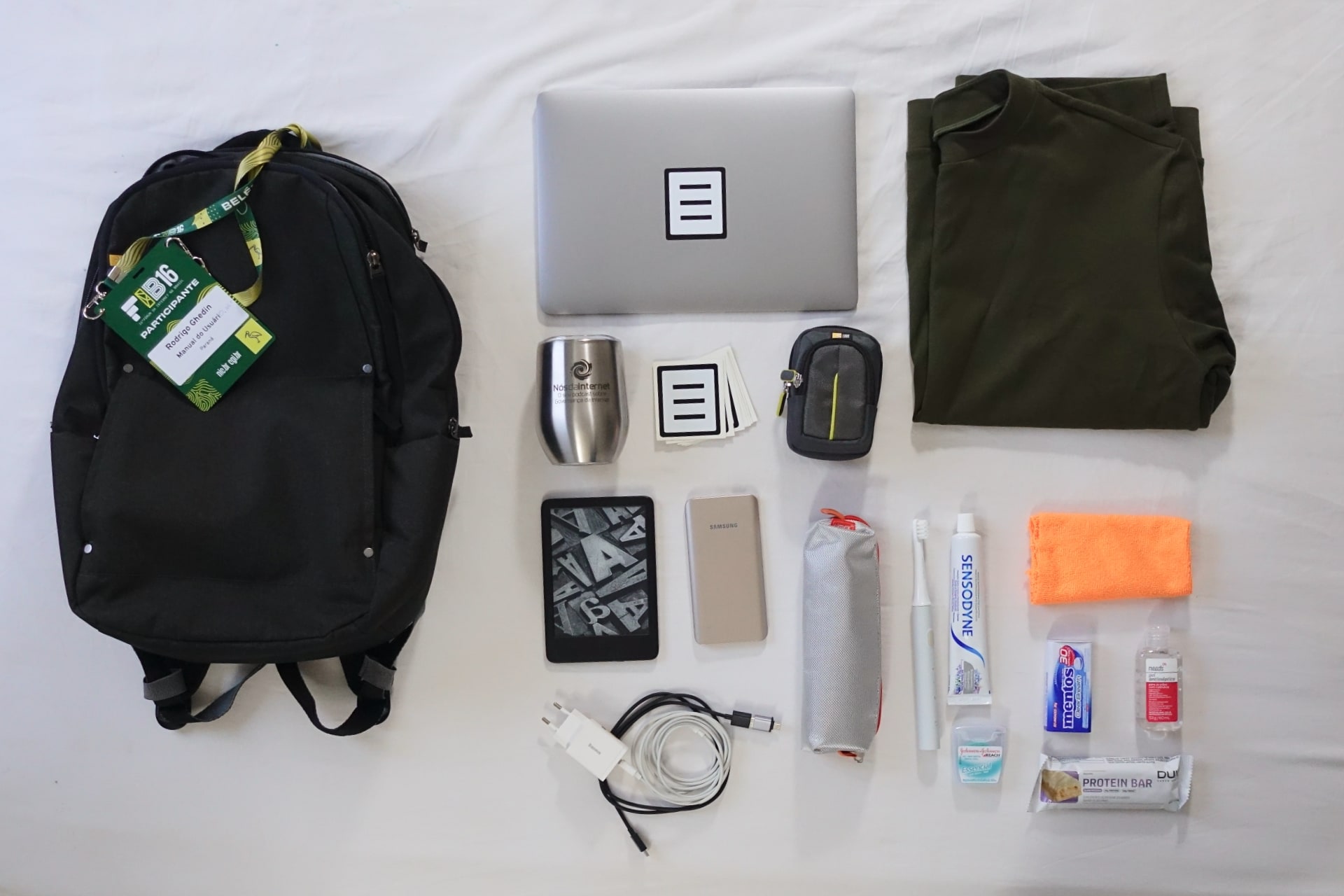 Foto de cima de uma mochila preta, com um crachá, e vários itens à direita, sobre um lençol branco.