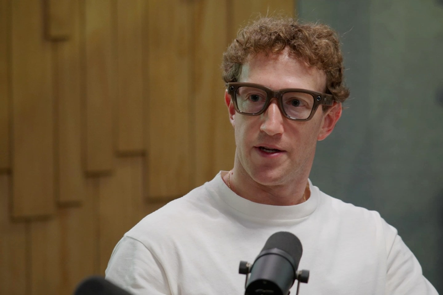 Mark Zuckerberg com óculos Meta Ray-Ban, camiseta branca e um microfone à sua frente.