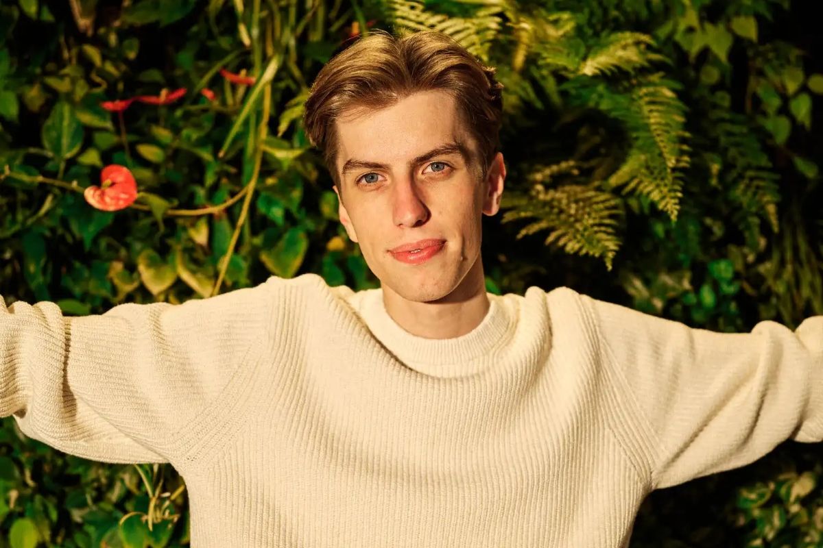 Homem branco, de cabelos claros e suéter branco, de braços abertos, à frente de muitas plantas.