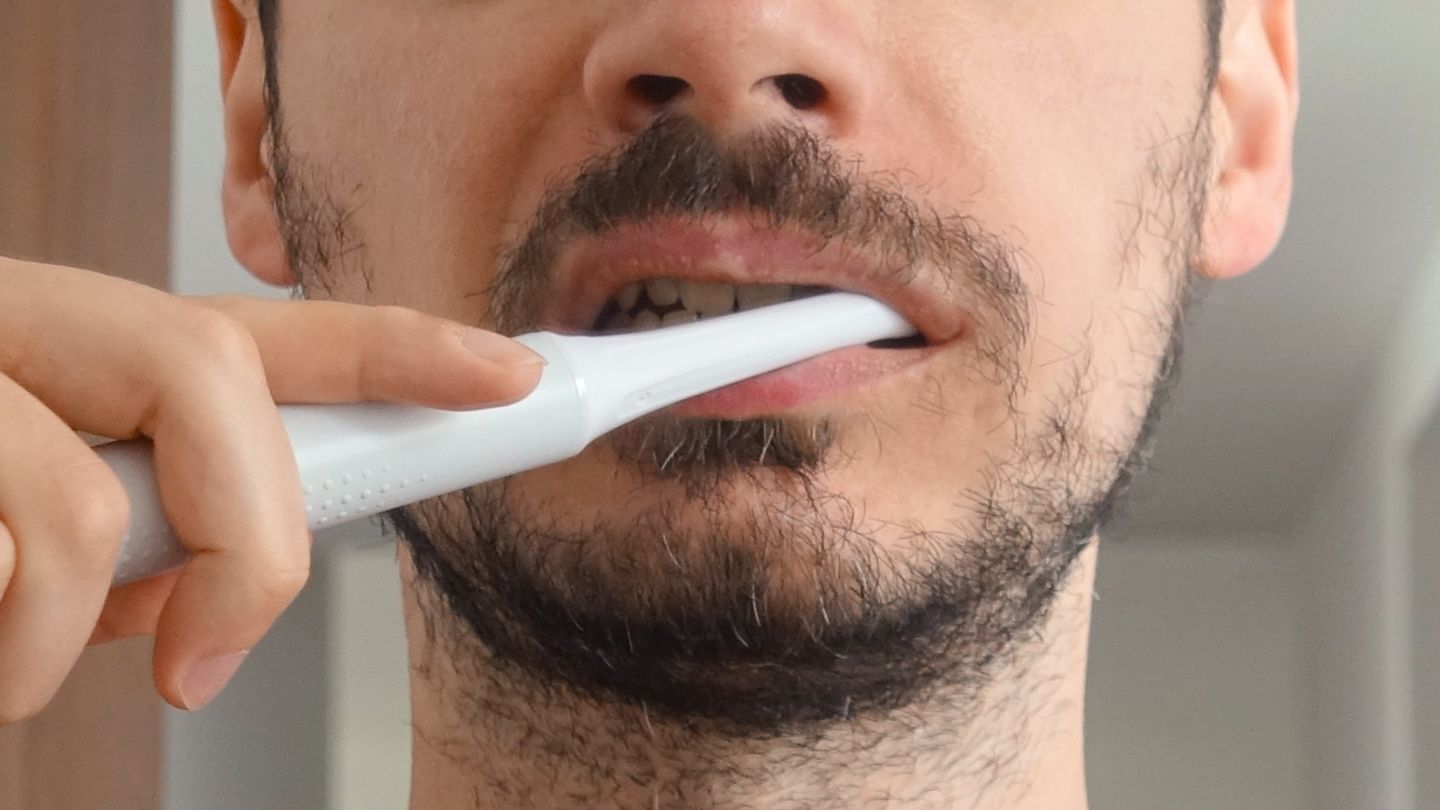 Homem escovando os dentes com uma escova de dentes elétrica branca. Foto cortada, do pescoço ao nariz.